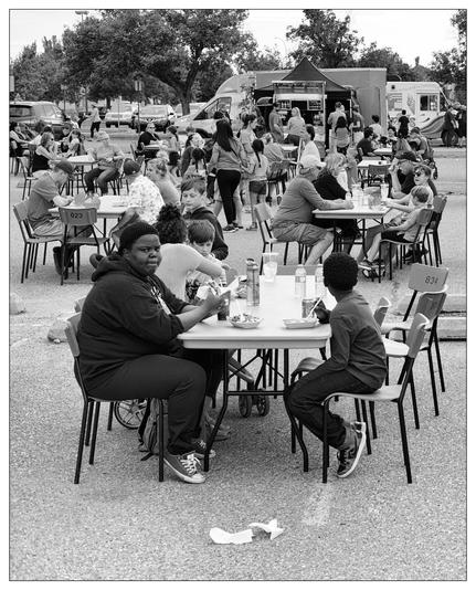 The image captures a vibrant outdoor scene at a food truck event. Numerous people of various ages are sitting at long tables, presumably enjoying meals and drinks. There are multiple food trucks positioned in the background, indicating where the food is coming from. In the foreground, a woman and a young boy are seated at a table, both engrossed in eating. The photograph is monochromatic, adding a timeless quality to the casual, communal dining atmosphere. Overall, the setting appears lively and social, with trees and parked cars in the distant background suggesting an urban park or similar public space hosting the event.