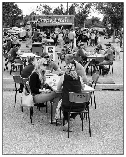 The image depicts an outdoor scene at a food event where multiple groups of people are seated at tables. At the center, there's a food truck named "Crepe Affair", indicating the type of food being served. Around the truck, several people are standing in line waiting to order. The people seated are engaged in conversations, enjoying their meals or drinks. The scene suggests a casual, social atmosphere typical of food truck gatherings or outdoor festivals. The photograph is in black and white, which adds a timeless quality to the image.