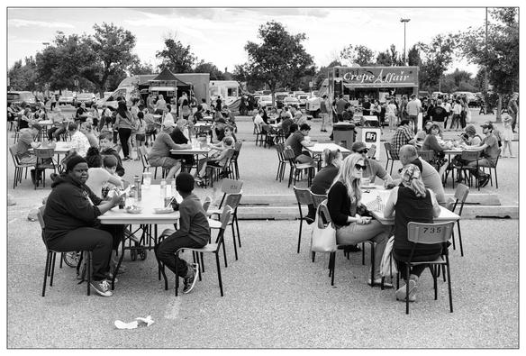 This image displays an outdoor gathering, possibly at a food truck event or an outdoor market. Many people are seated at tables enjoying meals and drinks. In the background, several food trucks are visible, including one named "Crepe Affair." It appears to be a lively community event, with attendees of various ages engaged in eating, talking, and socializing in what looks like a parking lot converted temporarily for this gathering. The scene is captured in black and white, adding a timeless feel to the photo.

This is the entire scene.  The previous two images were the isolated left and right hand portions.