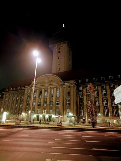 Das Bild zeigt ein großes, klassizistisches Gebäude bei Nacht, beleuchtet von Straßenlaternen. Es handelt sich um das Rathaus Spandau in Berlin. Das Gebäude verfügt über einen markanten Turm mit einer Uhr und einer Wetterfahne an der Spitze. Die Fassade ist mit Säulen und großen Fenstern geschmückt, und über dem Eingang befindet sich ein Zierbogen. Das Gebäude ist von einer Straße mit sichtbaren Fahrbahnmarkierungen umgeben, und auf dem Bürgersteig sind ein paar Menschen zu sehen. Vor dem Gebäude ist ein Baum mit kahlen Ästen zu sehen, und auf der rechten Seite des Bildes ist ein Straßenschild teilweise sichtbar. Der Himmel ist dunkel, was darauf hindeutet, dass es Nacht ist. Die gesamte Szene ist ruhig und gelassen, wobei sich das Gebäude vom Nachthimmel abhebt.