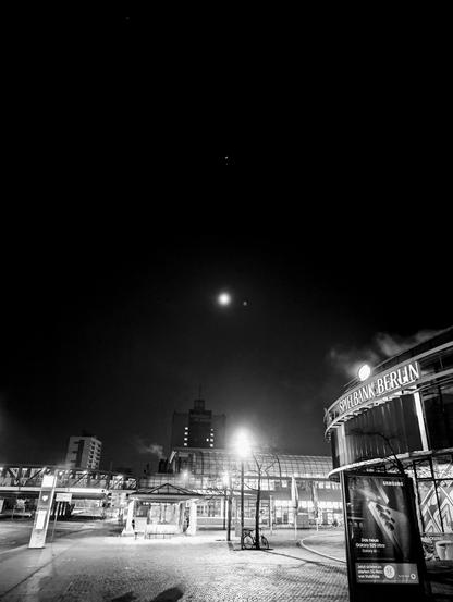 Das Bild zeigt eine nächtliche Szene am Bahnhof Rathaus Spandau in Berlin, Deutschland. Der Himmel ist dunkel, ein heller Mond und ein paar Sterne sind zu sehen. Das Bahnhofsgebäude ist gut beleuchtet, und das Schild "Spielbank BERLIN" ist deutlich sichtbar. Die Architektur weist eine moderne Glasfassade auf, und der Bahnhof ist von städtischen Elementen wie Straßenlaternen, einer Bushaltestelle und einer Fußgängerbrücke umgeben. Eine digitale Werbung für das Samsung Galaxy S21 Ultra ist auf einer Plakatwand zu sehen. Das Kopfsteinpflaster ist sichtbar, und in der Nähe ist ein Fahrrad geparkt. Die Gesamtatmosphäre ist ruhig und gelassen, mit minimaler Aktivität.