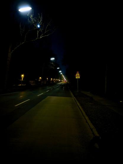 Das Bild zeigt eine nächtliche Szene auf einer ruhigen Straße. Die Straße wird von Straßenlaternen beleuchtet, die einen sanften Schein auf den Bürgersteig und die Umgebung werfen. Die Straße ist auf der linken Seite von Bäumen gesäumt, deren Äste sich vom dunklen Himmel abheben. Parallel zur Straße verläuft ein Fußgängerweg, der von einem Bordstein begrenzt wird. Auf der rechten Seite des Bildes ist ein dreieckiges Warnschild mit rotem Rand und gelbem Hintergrund zu sehen, das auf eine mögliche Gefahr hinweist. Das Schild ist mit einem schwarzen Ausrufezeichen versehen, das den Fahrer zur Vorsicht mahnt. Die Straße scheint nass zu sein und reflektiert das Licht der Straßenlaternen. In der Ferne ist der schwache Schein der Scheinwerfer eines Fahrzeugs zu sehen, was der Szene ein Gefühl von Tiefe verleiht. Die Gesamtatmosphäre ist ruhig und gelassen, wobei die Dunkelheit der Nacht mit dem künstlichen Licht kontrastiert.