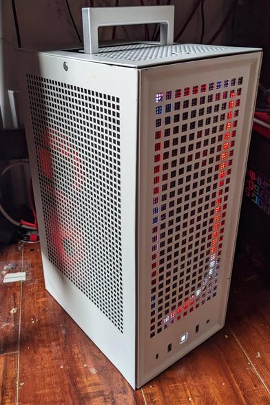 Three quarter view photo of a small, tall, white PC with red LED strips and fans illuminating it from the inside. It sits on a wood floor with it's silver carrying handle visible on top.