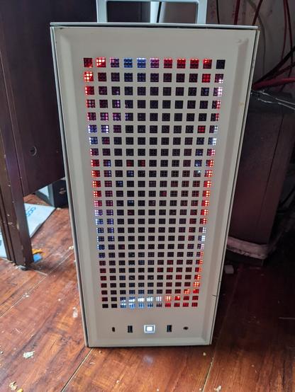 Front view photo of a small, tall, white PC with red LED strips and fans illuminating it from the inside. It sits on a wood floor with it's silver carrying handle visible on top.