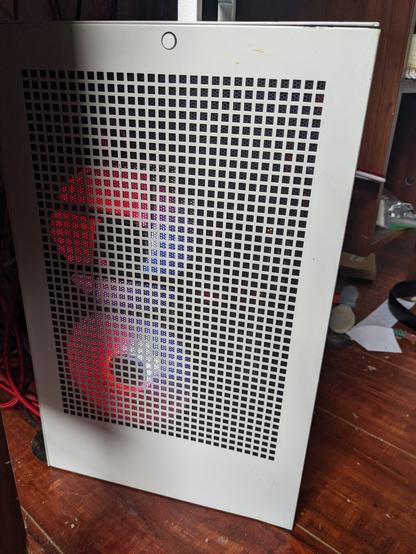 Side view photo of a small, tall, white PC with red LED strips and fans illuminating it from the inside. It sits on a wood floor with it's silver carrying handle visible on top.