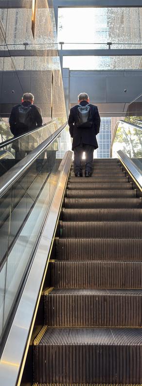 Vertical panorama of a man riding an upward escalator into the morning sun flooding the Marunouchi area in Tokyo, Japan.
