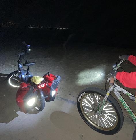 Two bikes paused on a night ride on the paved coastal trail above the Monterey Bay. At left, a silver-blue Tern NBD with red Ortleib panniers, whose reflective shapes are illuminated by the lights of the bike behind it, partly visible, at right, a Money Lynch Beast Mode Ripper.  #TernNBD #BeastMode