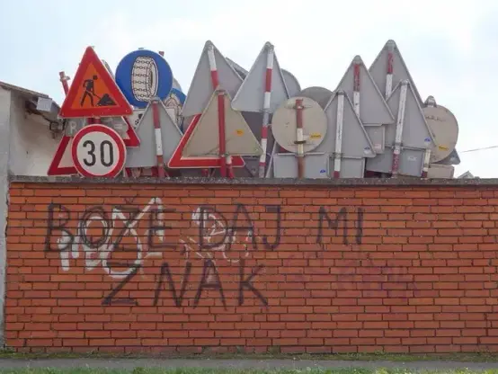 Text on the stone wall  is "Bože,daj mi znak!" translates to "God,give me a sign!", and there is bunch of road sign behind that stone wall. The irony is that text author didn't see signs asking for another one.