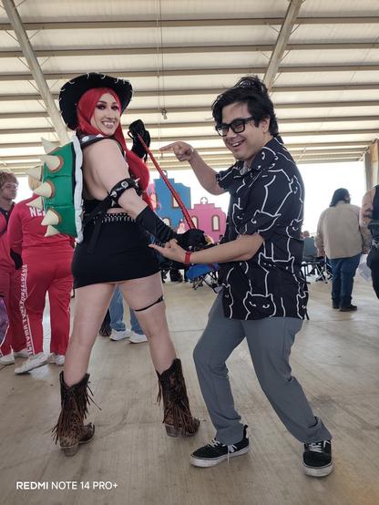 A close-up, angled shot of a Bowsette cowgirl cosplay and a person pointing to her in a black cat shirt, a 'REDMI NOTE 14 PRO+', visible in the lower left corner. The background is blurred.