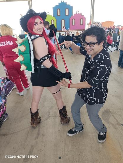 A close-up, angled shot of a Bowsette cowgirl cosplay and a person pointing to her in a black cat shirt, a 'REDMI NOTE 14 PRO+', visible in the lower left corner. The background is blurred.