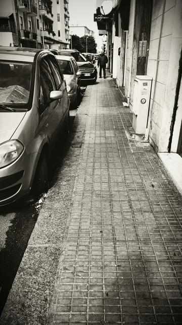 Un marciapiede stretto lungo una strada urbana, costeggiato da un lungo filare di auto parcheggiate in fila serrata, alcune con i finestrini coperti da riflessi o protezioni solari. L’asfalto è sconnesso ai margini, con foglie e detriti accumulati tra il bordo del marciapiede e le ruote delle vetture. Tracce di pioggia appena passata. Sulla destra, il muro degli edifici è segnato dal tempo, con insegne, citofoni e un’antenna parabolica che sporge. Un uomo in giacca imbottita cammina lontano, dando le spalle all’osservatore, diretto verso l’orizzonte. La fotografia in bianco e nero accentua il contrasto tra le superfici ruvide e lisce, creando un senso di profondità e di solitudine urbana.