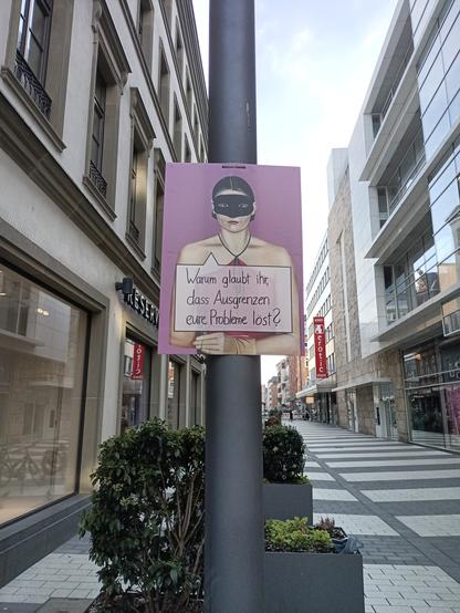 Ein Plakat an einem Mast in der Fußgängerzone in Mannheim. Auf dem Plakat ist eine Damie mit Augenmaske und der Schriftzug "Warum glaubt ihr das Ausgrenzung Eure Probleme löst?"