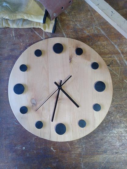 Une pendule ronde en bois ciré, faite de planche collées ensembles, avec des pastilles rondes et noires en relief pour marquer les heures.