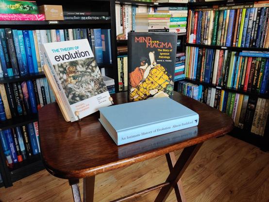 A photo of three books standing on a small, brown, wooden table. On the left, on a small wooden book stand, The Theory of Evolution, showing a small owl (a Ferruginous Pygmy-Owl) perched in a tree. In the middle, lying down, An Intimate History of Evolution, showing a teal-coloured book without its dustjacket. On the right, standing up, Mind Over Magma, showing a colourful photomicrograph of a Hedenbergite quartz syenite from the New Jersey Highlands (according to the book's dustjacket). The background shows black shelves full of books and part of the beech-coloured laminate floor.