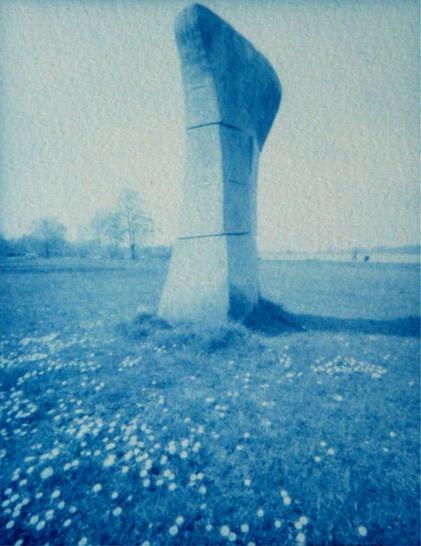 Eine blaue Fotografie zeigt einen bearbeiteten Stein vom Bildhauer Jesper Neergaard. Im Vordergrund sind Gänseblümchen im Gras abgebildet. Im Hintergrund eine Seenlandschaft, links ein paar Bäume.