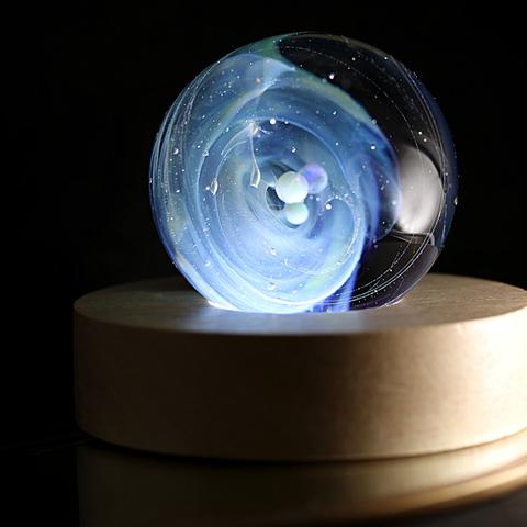 L'image montre une sphère en verre posée sur un socle circulaire en bois clair. La sphère est une œuvre soufflée qui contient un tourbillon de nuances bleutées et blanches, évoquant une galaxie en mouvement. De petites bulles de verre, semblables à des planètes en suspension, flottent en son centre et capte

La sphère est rétroéclairée, ce qui accentue la profondeur et la brillance des couleurs. Les reflets lumineux et les minuscules particules capturées dans le verre donnent l'illusion d'un espace infini. Le fond est sombre, ce qui met en valeur la lueur douce et presque mystique de l'objet. L'ensemble dégage une sensation de calme, de mystère et de fascination pour l'infiniment grand contenu dans un si petit un Univers en verre. 

The image shows a glass sphere placed on a circular light-colored wooden base. The sphere is a blown work containing a swirl of blue and white hues, evoking a moving galaxy. Small glass bubbles, like suspended planets, float in its center and capture

The sphere is backlit, which accentuates the depth and brilliance of the colors. The light reflections and tiny particles captured in the glass give the illusion of infinite space. The background is dark, highlighting the object's soft, almost mystical glow. The whole thing exudes a sense of calm, mystery, and fascination for the infinitely vast universe contained within such a small glass universe.