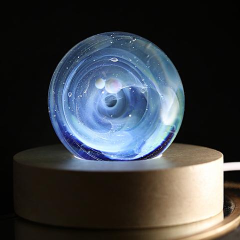 L'image montre une sphère en verre posée sur un socle circulaire en bois clair. La sphère est une œuvre soufflée qui contient un tourbillon de nuances bleutées et blanches, évoquant une galaxie en mouvement. De petites bulles de verre, semblables à des planètes en suspension, flottent en son centre et capte

La sphère est rétroéclairée, ce qui accentue la profondeur et la brillance des couleurs. Les reflets lumineux et les minuscules particules capturées dans le verre donnent l'illusion d'un espace infini. Le fond est sombre, ce qui met en valeur la lueur douce et presque mystique de l'objet. L'ensemble dégage une sensation de calme, de mystère et de fascination pour l'infiniment grand contenu dans un si petit un Univers en verre. 

The image shows a glass sphere placed on a circular light-colored wooden base. The sphere is a blown work containing a swirl of blue and white hues, evoking a moving galaxy. Small glass bubbles, like suspended planets, float in its center and capture

The sphere is backlit, which accentuates the depth and brilliance of the colors. The light reflections and tiny particles captured in the glass give the illusion of infinite space. The background is dark, highlighting the object's soft, almost mystical glow. The whole thing exudes a sense of calm, mystery, and fascination for the infinitely vast universe contained within such a small glass universe.