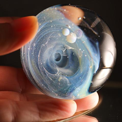 Une main tient délicatement une sphère en verre, mettant en valeur ses détails fascinants. La sphère est une œuvre soufflée qui semble capturer un petit univers en son cœur. On y distingue un tourbillon de nuances bleues et blanches, créant une illusion de profondeur, comme une galaxie en formation.

De petites bulles translucides flottent à l’intérieur, reflétant la lumière et ressemblant à de minuscules planètes suspendues dans l’espace. Des particules scintillantes sont dispersées dans le verre, ajoutant un effet étoilé.

La lumière éclaire la sphère, révélant ses reflets et ses détails subtils. L’arrière-plan est sombre, ce qui accentue la luminosité et la transparence de l’objet. La présence des doigts autour de la sphère donne une échelle, soulignant la finesse et la délicatesse de cette pièce unique.



A hand delicately holds a glass sphere, highlighting its fascinating details. The sphere is a blown work that seems to capture a small universe within its center. A swirl of blue and white hues can be seen, creating an illusion of depth, like a galaxy in formation.