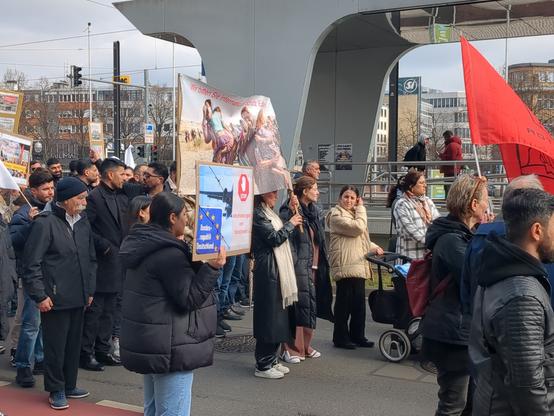 Demonstration von Jesidinnen und Jesiden für ihr Bleiberecht. Rote Flagge. Menschen mit Schildern und Parolen