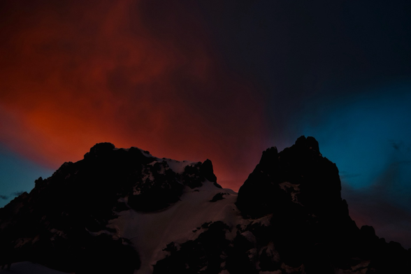 A partially snow covered rocky portion of Mt. Hood at dusk with red, sunlit clouds in a medium blue sky.