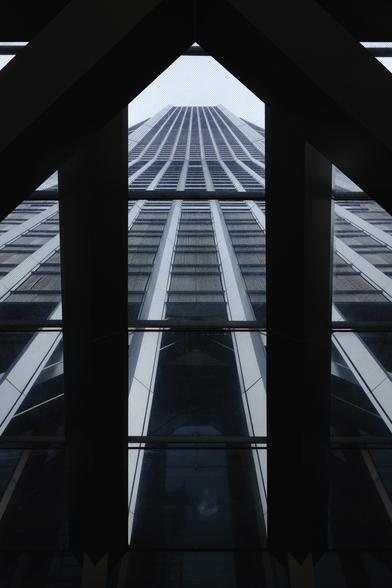 An upwards photo of a very tall building from beneath a glass sealing. The building itself has long stretched vertical lines up to the top. It’s grey in tone. The glass sealing has some dark bars which frame the whole image