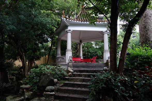 A tranquil scene unfolds in a lush garden, featuring a traditional Chinese-style pavilion as its focal point. The pavilion is elevated on a stone platform, accessible by a set of weathered stone steps leading up from the foreground. The structure boasts a grey tiled roof with ornate, upturned corners and decorative ridge details. It is supported by four round, light-coloured stone pillars.
Inside the pavilion, two vibrant red, ornate benches face each other, inviting rest and contemplation. The platform of the pavilion is surrounded by a low, decorative stone railing.
The garden is abundant with mature trees and dense greenery, creating a sense of enclosure and shade. Various types of foliage, from leafy shrubs to larger trees with textured bark, fill the frame, suggesting a well-established and cared-for space. The lighting is soft and diffused, typical of a shaded garden environment. The overall atmosphere is one of peace and serenity.