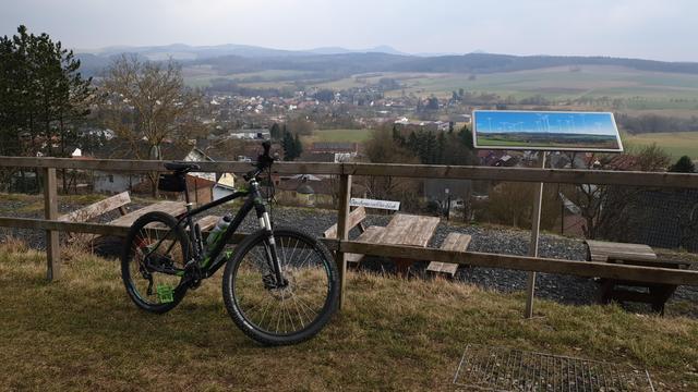 Im Vordergrund ist ein Fahrrad an einen Zaun gelehnt. Dahinter ist ein Rastplatz mit Bänken und Tischen zu erkennen. Der Blick geht dann über das Dorf Hofaschenbach zu den Bergen der Rhön.