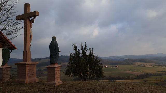 Auf der linken Bildseite ist eine Kreuzigungsgruppe zu ernennen. Ganz links sieht man noch ein Stück von einer Kapelle. Die Kreuzigungsgruppe und die Kapelle stehen auf einer Anhöhe, von der man über das Tal zu den höheren Bergen der Rhön blickt.
