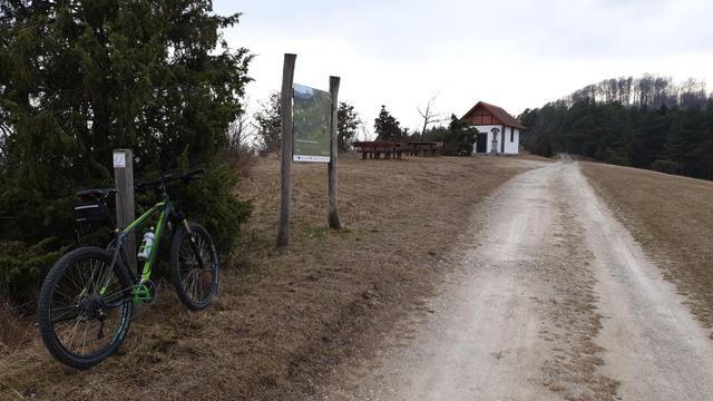 Auf der rechten Bildseite führt ein Feldweg auf einen Berg zu. Links ist ein Fahrrad an einen Pfosten mit einem Wanderweg-Zeichen angelehnt, einem "U" für die Extratour Ulmenstein. Etwas weiter steht eine Tafel mit Erklärungen zu der Landschaft. Weiter hinten ist ein kleiner Rastplatz mit Tischen und Bänken und dahinter eine Kapelle.