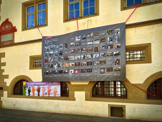 Ein großes Banner zeigt bemerkenswerte Pressefotos, die nach Jahrzehnten von den 1950er bis 2020er Jahren sortiert sind und an der Wand eines Gebäudes hängen. Unten ist ein kleineres Banner, das für den „Tag gegen Rassismus“ wirbt.

A large banner displays notable press photographs organized by decade from the 1950s to the 2020s, hanging on the wall of a building. Below, a smaller banner promotes „Tag gegen Rassismus“ (Day Against Racism).