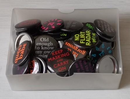 Little boxful of maybe 50 or so button badges.  They're black with coloured writing, and they're jumbled together so that not all the writing can be seen.  Slogans of the ones on the top layer include
• Keep masking and carry on
• No flirt radar
• Old enough to know my own mind
• Fat & gorgeous
• Queer babe.
A few are upside down, showing silvery metal backs.  They don't have their pins yet.  
Each badge is one inch in diameter, although that might not be obvious from the picture.