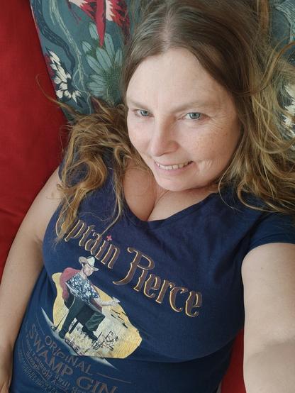 A white woman in her fifties lying on a red sofa, wearing a dark blue t-shirt with a print of Hawkeye Pierce from the TV- series MASH.. It is a fake advert for his swamp distilled gin.