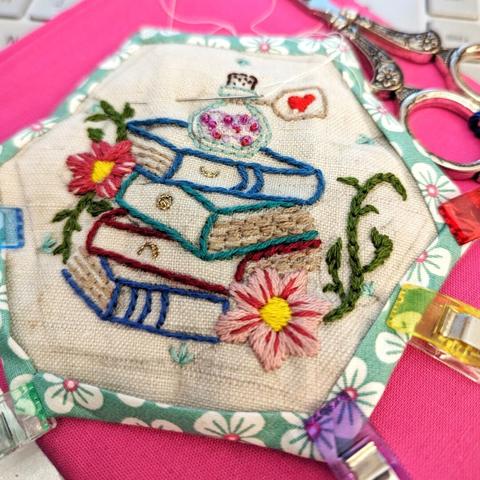 On the fabric lies a linnen hexagon with the embroidery of witchy books and some flowers. The hexagon is in the process to be framed by another fabric by hand, the needle with a thin white thread rests in the fabric.