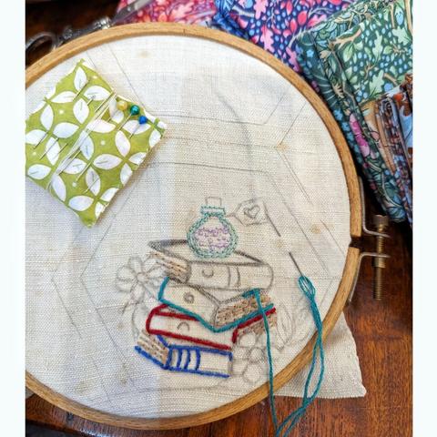 Embroidery hoop with old linnen. Inside a painted hexagon is an embroidered stack of old books, a little bottle and 2 flowers and some tendrils. A green-white pincushion and a needle with turquoise embroidery thread are lying on the fabric.