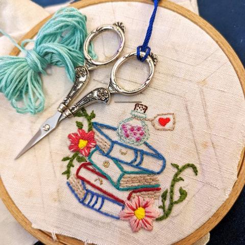 Embroidery hoop with old linnen. Inside a painted hexagon is an embroidered stack of old books, a little bottle and 2 flowers and some tendrils. Silver scissors and turquoise embroidery thread are lying on the fabric.