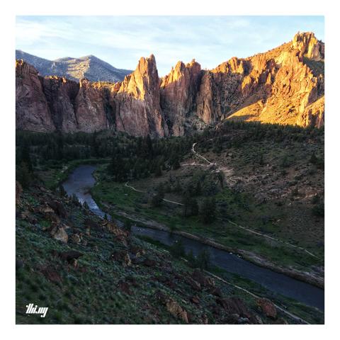 Photo of a partially forested valley with a river bend (Crooked River), surrounded by orange rocky cliffs during sunset. A small path is winding up the hills on the other side of the river. Slightly overcast, pale blue sky.