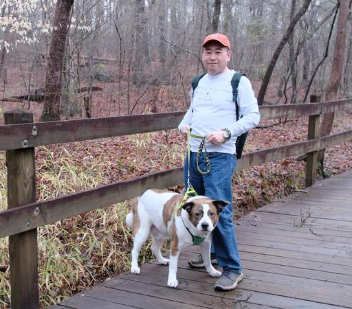 A man with an orange cap standing on a wooden bridge over a stream holding his white and brown dog at his feet, staring at the camera