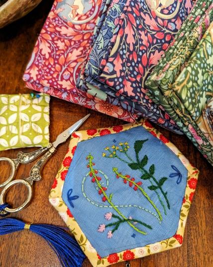 Brown table, finished, big cloth hexagon out of blue linnen, embroidered with wild flowers. A silver scissor, green-whit pincushion and small packs of beautiful, fabric with lots of flowers lying alongside.