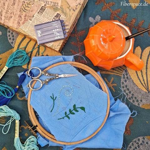 Petrol blanket with weaved in flowers and birds. on the blanket: a wooden embroidery hoop with some blue linnen inside. There is a painted on pattern with a hexagon and some widl flowers, some are embroidered, the rest not. Silver scissors, a needle case, embroidery thread, an orange pumpkin mug with coffee and a paper box are scattered around.