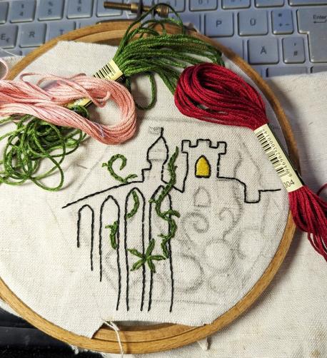 Embroidery hoop with old linnen. Inside a painted hexagon you can see the outlines (drawn with a pencil) of a castle, a bridge and tendrils and flowers - which are partially embroidered. Green, pink and dark red embroidery thread are lying on the fabric.