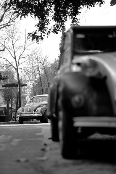 Black-and-white photo of a vintage, mid-1960s Volkswagen beetle, peeking around the corner from behind a vintage Citroën 2CV.