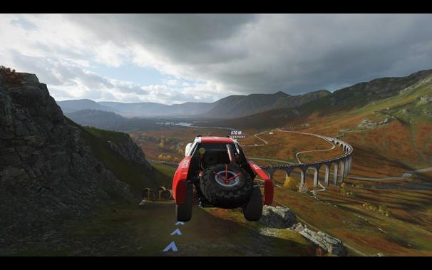 Alt Text Description: A first-person perspective of a red off-road vehicle, likely an SUV or truck, driving on a narrow mountain road during daylight. The terrain is rocky and the sky is cloudy, with a river visible down below. There are no UI elements in view.