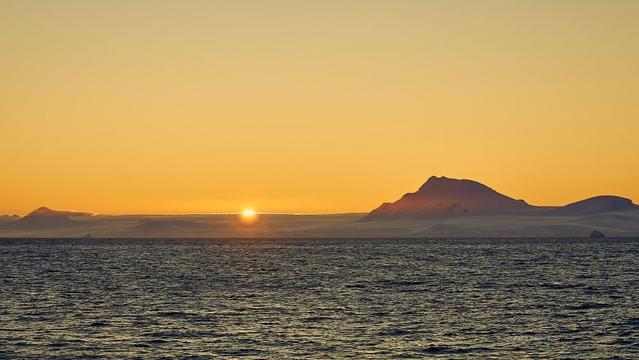 Setting sun lighting the skies up in bright yellow. There's a haze floating over the snow-covered, and backlit Antarctic landscape.  There's a larger cruise ship in the shadows on the right, mid-frame.  The seas have a light chop, still calm enough to provide sharpness with a 1/250th second shutter as we sail past slowly.