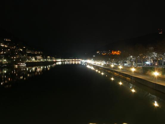 Eine Langzeitbelichtung vom Neckar in Heidelberg. Man seiht den Fluß, die beleuchteten Straßen rechts und LInks, die Gebäude, die die Berge hinauf gehen und weiter hinten eine weitere Brücke. Etwa in der Mitte ist das erleuchtete Schloß erkennbar.