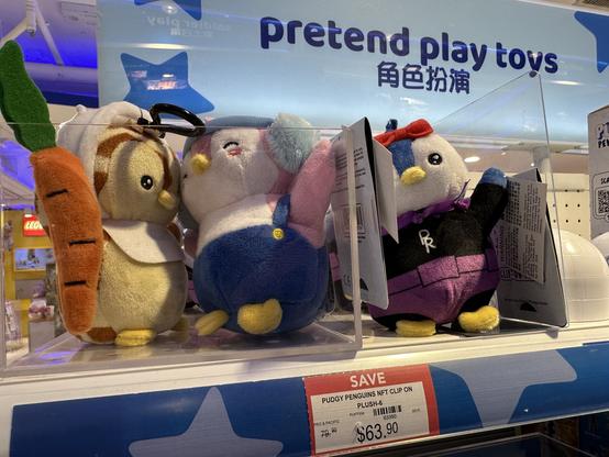 Drei komische Plüschtiere liegen im Regal. Auf dem Preisschild unter dem Produkt steht „PUDGY PENGUINS NFT CLIP ON PLUSH-6“ über den Tiere die Marke „pretend play toys“