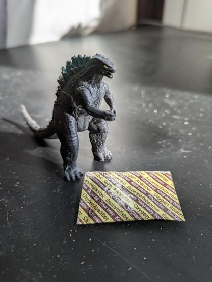 The same sachet from the previous photo along with a tiny Monsterverse Godzilla on a black table.