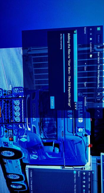 Photograph of an IPS LED panel displaying a file compression process. The main window is titled "Archive Manager" and shows the progress of adding files to a tgz file named "Star Wars - The Old Republic.tar.gz." The window indicates that 46 files remain to be added. The background has a Scania truck in a blue-toned image of a gorgeous blue truck with the word "SCANIA" visible on its side, parked in front of a massive building. The truck has a robust design with multiple lights and a mesh guard on the front.

The photograph was composed using a very simple small sensor camera driven by an Android operating system, shot in Total Darkness with the camera in full automatic mode, driven by Open Camera