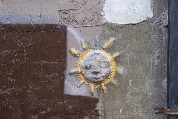 This sun medallion is an odd decorative part of a building exterior back wall patched many times with new cement and paint. It is raised up cement like a medallion that has been a bit eroded and rubbed off partly.  The yellow paint highlights some of the rays and the sphere is encircled with yellow paint partly over white outline paint.  There is a bit of medium gray-blue paint on the most raised part of the sun face.  The rest of the wall is textured and patched cement showing beige, white, brown and blue-gray sections as for an abstract painting.