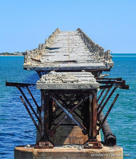 Crumbling highway atop rusting railroad trestle with ocean in the background.