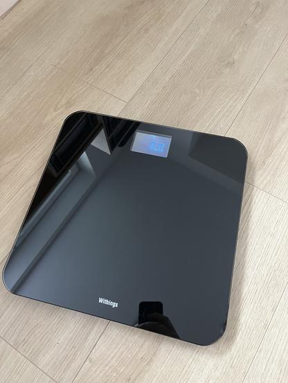 Eine moderne digitale Personenwaage mit einer glatten schwarzen Glasoberfläche und einem klaren Display mit „0,0 kg“. Die Waage liegt auf einem hellen Holzboden. Der Markenname „Withings“ ist unten zu sehen.